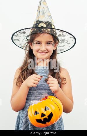 Pronta a fare il trick-or-treating. Una bambina che indossa un cappello di strega mentre tiene un jack o lanterna su uno sfondo bianco. Foto Stock