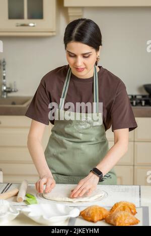 Una giovane casalinga in un grembiule verde prepara torte fatte in casa in una cucina luminosa. Foto Stock