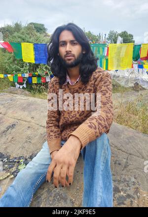 Un giovane uomo con capelli lunghi e barba che guarda la macchina fotografica mentre si siede sulla roccia con lo sfondo delle bandiere tibetane di preghiera a Rewalsar Foto Stock