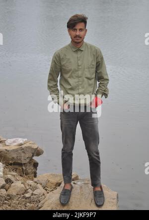 Tutta la lunghezza di un ragazzo che guarda la macchina fotografica, posando con le mani in tasca mentre si alza sulla pietra vicino al lago Foto Stock