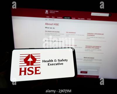 Persona che tiene uno smartphone con il logo dell'agenzia britannica Health and Safety Executive (HSE) sullo schermo davanti al sito Web. Mettere a fuoco sul display del telefono. Foto Stock