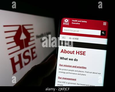 Persona che tiene il cellulare con il sito web dell'agenzia britannica Health and Safety Executive (HSE) sullo schermo di fronte al logo. Concentrarsi sul centro del display del telefono. Foto Stock