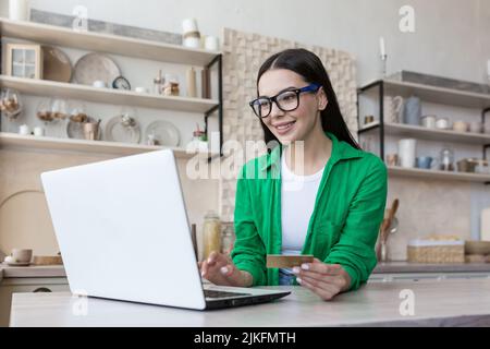 Giovane bella donna a casa in cucina in bicchieri e una camicia verde fa acquisti online in un negozio online, brunette utilizza un computer portatile e tiene una carta di credito in mano Foto Stock