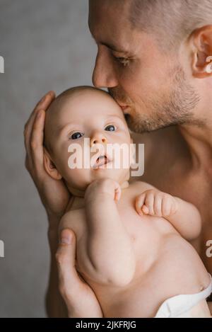 Un giovane padre tiene il figlio neonato in un pannolino tra le braccia e lo bacia Foto Stock