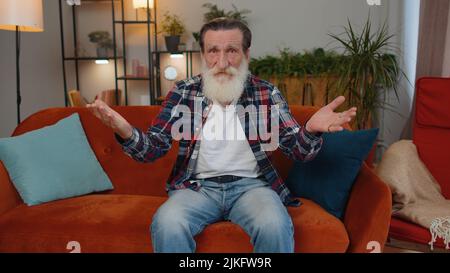 Che cosa. Perché. Anziano nonno alzando le mani in espressione indignata, chiedendo motivo di fallimento, dimostrando irritazione incredulità da problemi in casa appartamento. Ragazzo anziano in camera seduta sul divano Foto Stock