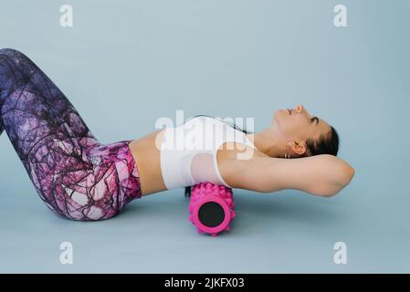 Una donna esegue il rilassamento miofasciale dei muscoli iperflessibili della schiena con un rullo massaggiante su sfondo blu. Il concetto di prevenzione Foto Stock