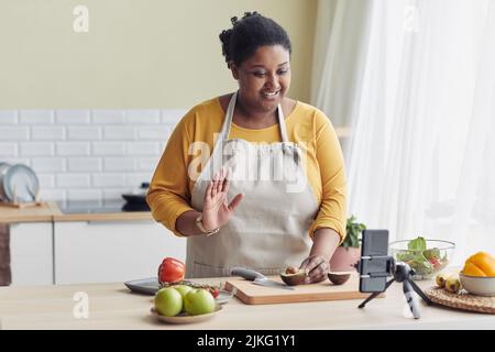 Ritratto di giovane donna nera che cucinano pasto sano in cucina e ondeggiando alla macchina fotografica durante il livestream, spazio di copia Foto Stock