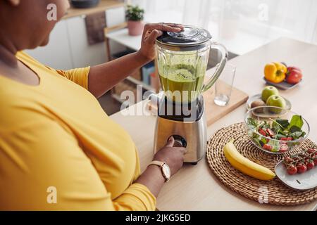 Closeup ad angolo elevato di donna nera sovrappeso utilizzando il frullatore mentre si fa un frullato sano in cucina a casa Foto Stock