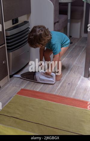 Il bambino, simpatico e diligente, pulisce la roulotte con un aspirapolvere. Foto Stock