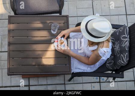 Una donna solitaria con un cappello bianco si siede in un caffè all'aperto da solo e beve caffè in una giornata estiva. Primo piano, vista dall'alto Foto Stock