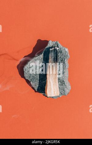 Fuoco palo santo bastone con fumo su pietra stand su sfondo arancione rosso, incenso ardente. Composizione verticale con bastoncini di legno santo per meditat Foto Stock