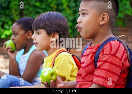 bambini multietnici con zaini seduti in strada all'ingresso della scuola mangiando mele. Concetto di ritorno a scuola. Gruppo di bambini multietnici Foto Stock