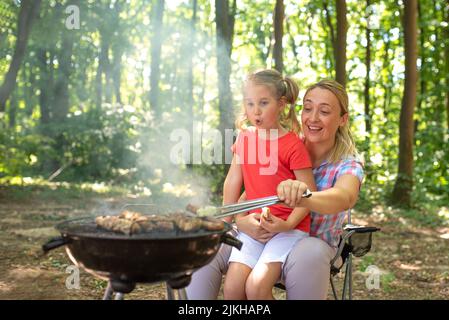 Una giovane madre felice campeggio e cucina barbecue in una foresta con sua figlia Foto Stock