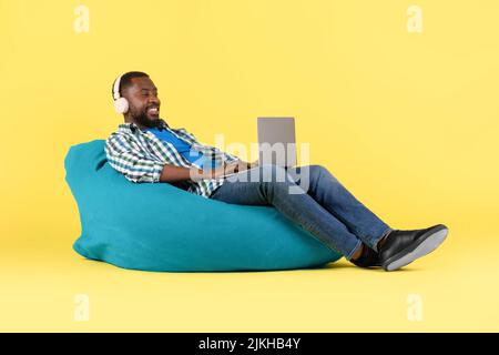Uomo nero che usa il laptop che indossa le cuffie seduta su sfondo giallo Foto Stock