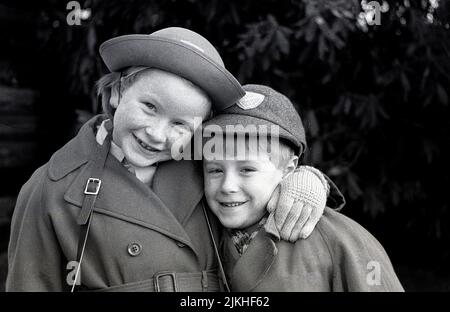 1963, storico, in piedi insieme fuori nella loro uniforme scolastica, con cappotti e indossare i loro cappelli scolastici, un abbraccio amorevole da sorella a suo fratello minore, Inghilterra. REGNO UNITO. Foto Stock