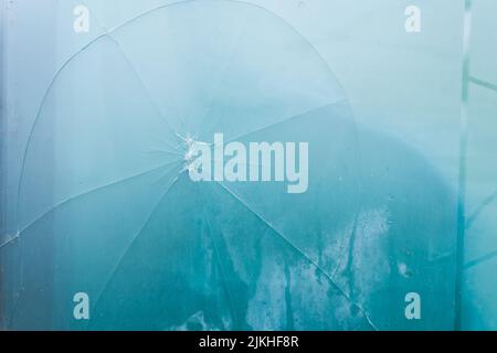 Un primo piano di un vetro rotto fessurato con un foro su sfondo nebbia blu Foto Stock