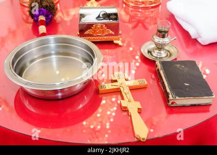 Una croce d'oro, la bibbia, una ciotola d'acqua Santa, e una scrigno su una tavola rossa della chiesa - cerimonia del battesimo Foto Stock
