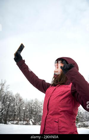 Un colpo verticale di una allegra donna caucasica in abiti rosa che prende selfie sotto la neve Foto Stock