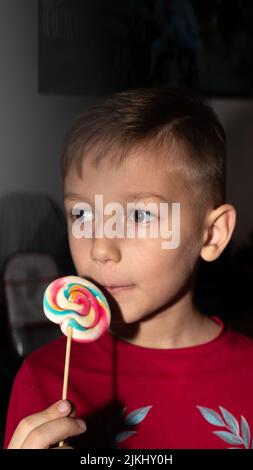 un ragazzo mangia una dolce caramella multicolore su un bastone, un ragazzo con un lecca di zucchero, che gli tiene in mano Foto Stock