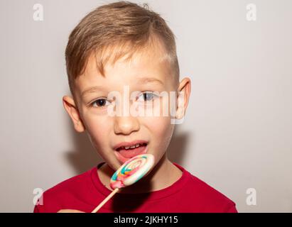 un ragazzo mangia una dolce caramella multicolore su un bastone, un ragazzo con un lecca di zucchero, che gli tiene in mano Foto Stock