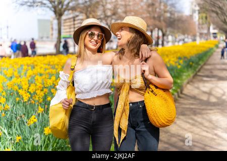 Amici in città con cappelli di paglia che camminano e sorridono accanto ad alcuni bei fiori gialli, godendo la primavera Foto Stock