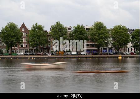 Due barche in rapido movimento che si muovono lungo il fiume Amstel sullo sfondo degli edifici di Amsterdam, nei Paesi Bassi. Foto Stock