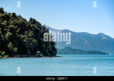 Un bellissimo paesaggio con molo in legno in acqua, verdi alberi di lievito e montagne contro il cielo blu alla luce del sole Foto Stock