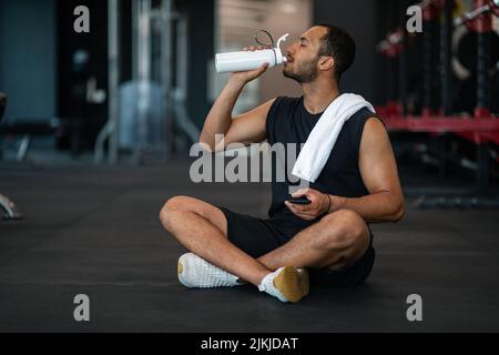 Giovane atleta maschio Nero bere acqua mentre si rilassa dopo l'allenamento in palestra Foto Stock