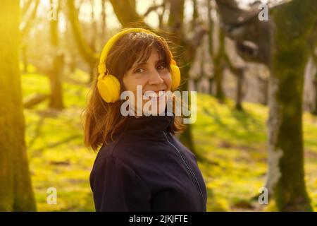 Una ragazza che ascolta la musica con le cuffie gialle nella foresta al tramonto, guardando la macchina fotografica Foto Stock