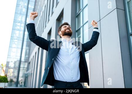 Allegro attraente giovane uomo d'affari caucasico con barba in causa solleva le mani e si rallegra per la vittoria Foto Stock