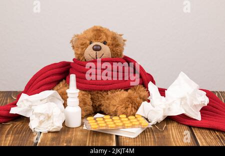 Un soffice orsacchiotto con spray nasale, pillole, sciarpa rossa e tessuti su una superficie di legno - affetti da malattia Foto Stock