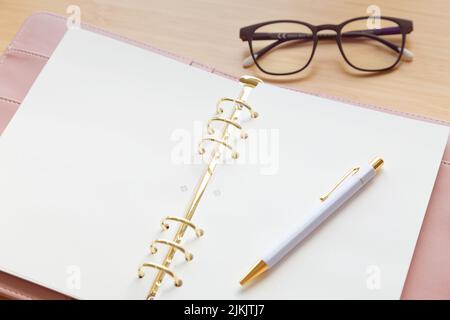 Un raccoglitore aperto color rosa pastello con penna e bicchieri su una scrivania di legno. Foto Stock