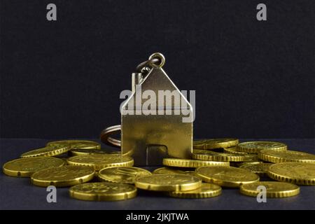 un primo piano di una piccola casa di metallo e monete sparse sullo sfondo nero. Foto Stock