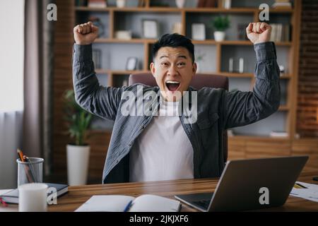 Felice eccitato uomo d'affari asiatico adulto con bocca aperta alza le mani in su, facendo il gesto di vittoria sul posto di lavoro Foto Stock