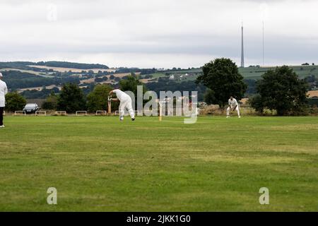Battitore gioca un colpo difensivo in avanti in una partita di cricket locale con Emley Moor Transmission Mast sullo sfondo nello Yorkshire Foto Stock