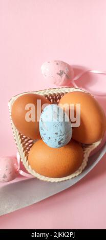 Una foto dall'alto di uova di pollo in una ciotola, le decorazioni rosa e blu dell'uovo di pasqua e una striscia grigia su uno sfondo rosa. Foto Stock