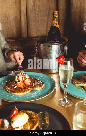 Un primo piano di mani che tagliano la frittella con la guarnizione del gelato con un secchio di champagne dietro Foto Stock
