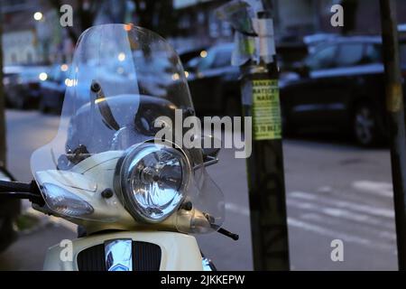 Un primo piano di uno scooter vespa d'epoca parcheggiato in strada al tramonto con una fila di auto parcheggiate sullo sfondo sfocato Foto Stock