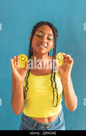 Una verticale di una giovane ragazza allegra di razza mista che tiene kiwi contro lo sfondo blu Foto Stock