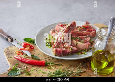 Piatto con braciole di agnello marinato crudo su tagliere di legno con olio e peperoncino su sfondo grigio in cucina Foto Stock