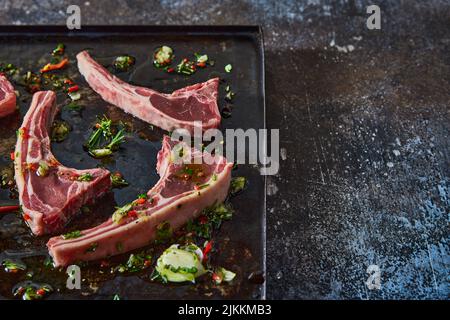 Da sopra di costolette di agnello non cotte poste su vassoio nero con verdure tritate e olio su tavola scura in cucina chiara Foto Stock