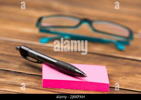 Un primo piano di una penna su un cartellino rosa appiccicoso con bicchieri blu sullo sfondo sfocato su un tavolo di legno Foto Stock