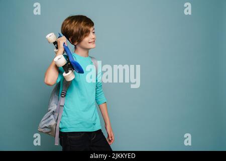 Ragazzo bianco zenzero sorridente mentre si posa con skateboard isolato sopra la parete blu Foto Stock