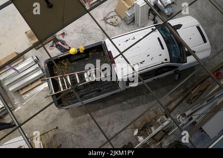 Il lavoratore di appaltatore caucasico nei suoi 30 anni e il suo moderno camion bianco di pick-up in un cantiere. Vista dall'alto sul soffitto. Tema dell'industria delle costruzioni. Foto Stock