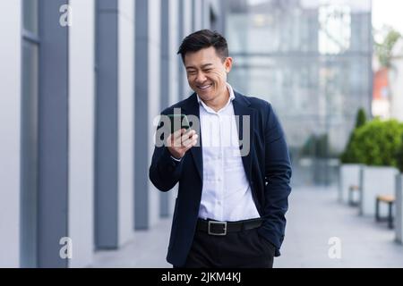 uomo asiatico freelance a piedi vicino business center tenere telefono, sorridente leggere notizie, uomo d'affari di successo Foto Stock