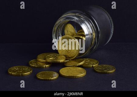 Un primo piano di monete Euro sparse e all'interno di un vaso di vetro su una superficie e sfondo nero. Foto Stock