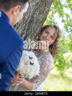 Un colpo verticale di sposi caucasici che si guardano l'un l'altro mentre si levano in piedi accanto ad un albero Foto Stock
