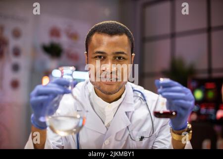Yong scienziato maschile africano che lavora con provette che indossano camice da laboratorio mentre esamina il campione di biochimica in provette e strumenti scientifici. Foto Stock