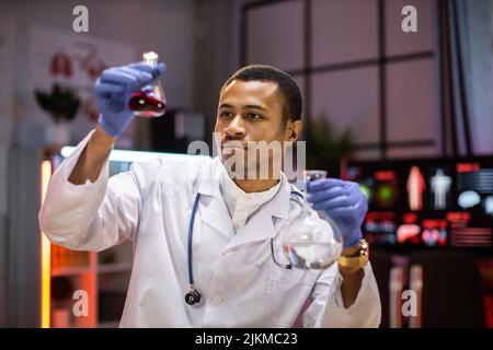 Yong scienziato maschile africano che lavora con provette che indossano camice da laboratorio mentre esamina il campione di biochimica in provette e strumenti scientifici. Foto Stock