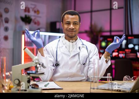 Yong scienziato maschile afroamericano rilassamento pratica yoga indossando camice da laboratorio lavorando in laboratorio mentre si esamina campione di biochimica in provetta e strumenti scientifici. Foto Stock